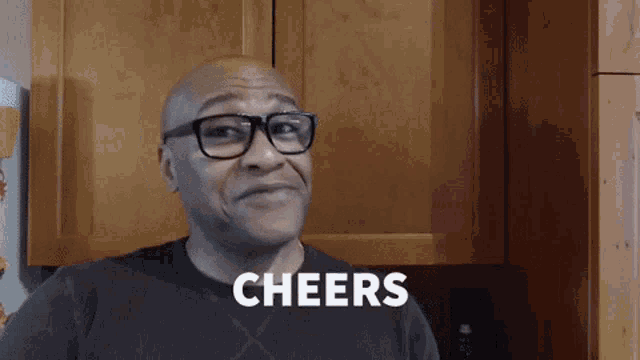 a bald man wearing glasses says cheers in front of a wooden cabinet