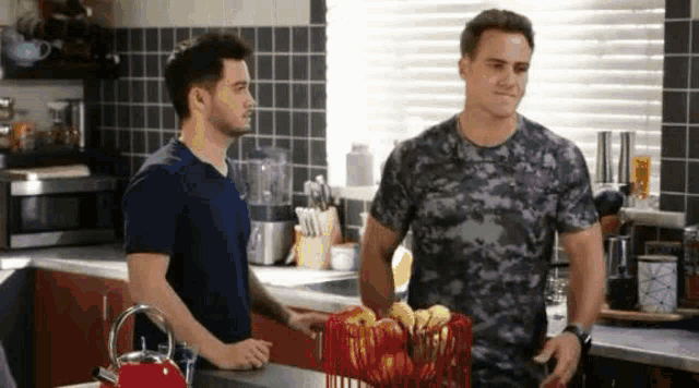 two men are standing in a kitchen talking to each other . one of the men is holding a basket of apples .