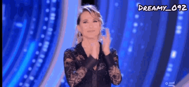 a woman in a black lace dress is clapping her hands on a blue background with dreamy_092 written on the bottom