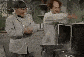 two men are standing next to each other in a kitchen . one of the men is dancing in front of an oven .