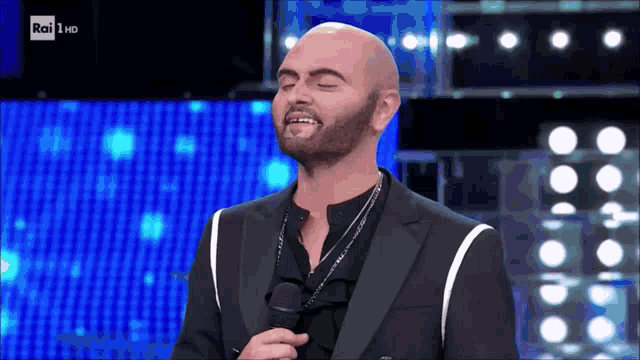 a bald man with a beard is singing into a microphone with rai hd in the background