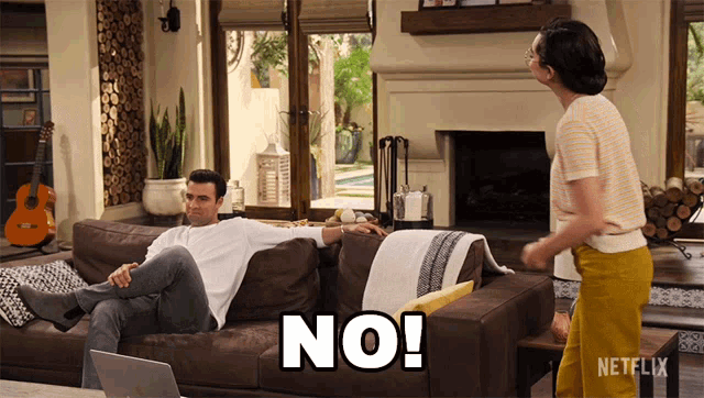 a man sits on a couch while a woman stands next to him and says " no "