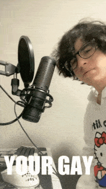 a man wearing glasses and a hello kitty shirt stands in front of a microphone with the words " your gay " written below him