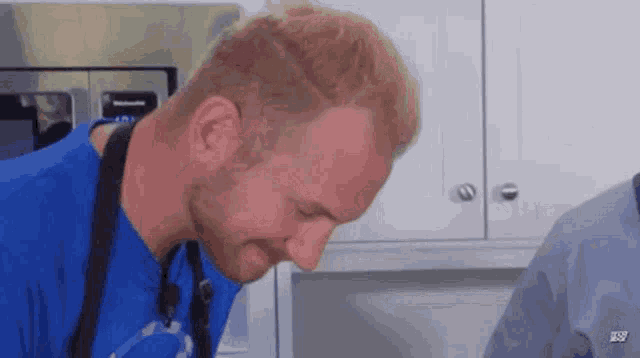 a man in a blue shirt and apron is standing in a kitchen looking down .