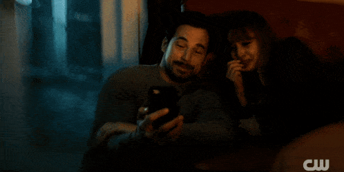 a man and a woman sit at a table looking at a cell phone with a cw logo in the background