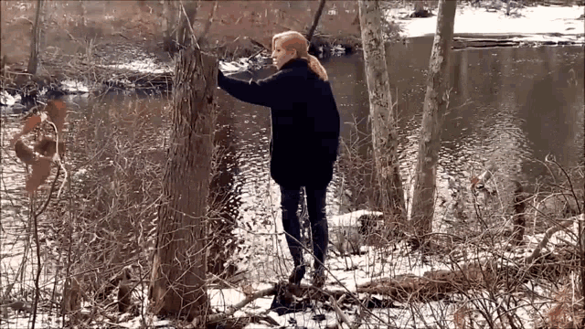 a woman in a black coat is standing in the woods near a river .