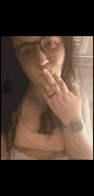 a woman wearing glasses is covering her mouth with her hands .