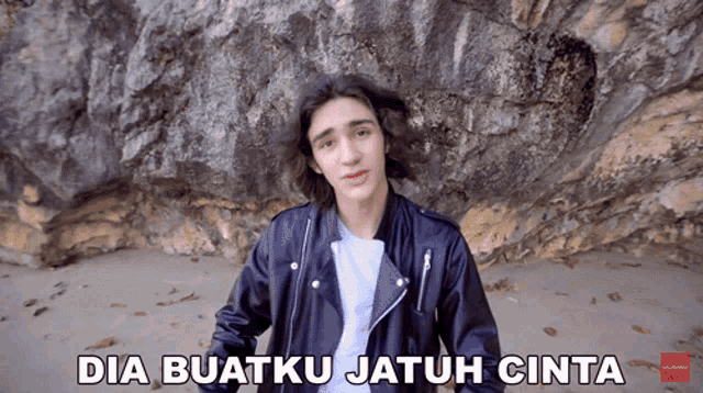 a young man in a black leather jacket stands in front of a rock wall with dia buatku jatuh cinta written on the bottom