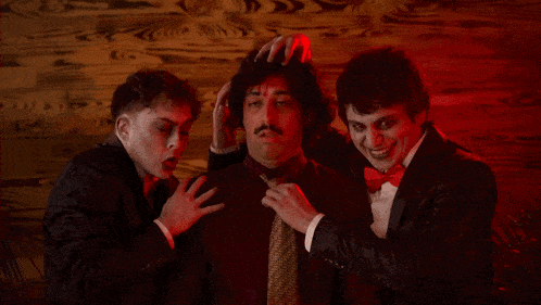 a man in a suit and tie is surrounded by two other men in tuxedos