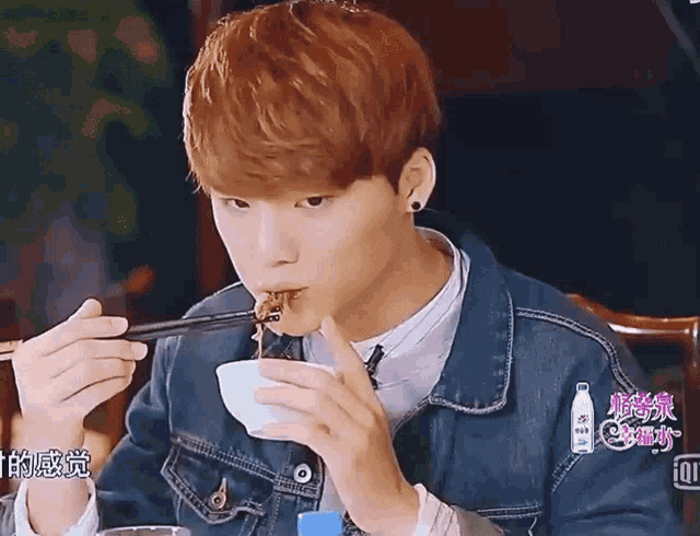 a man in a denim jacket is eating from a bowl with chopsticks