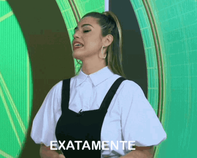 a woman in a white shirt and black overalls stands in front of a green background that says exactamente on it