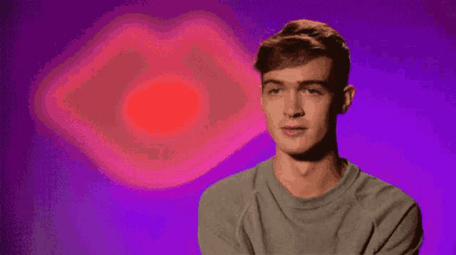 a man in a grey shirt is standing in front of a purple background with a red heart in the background .