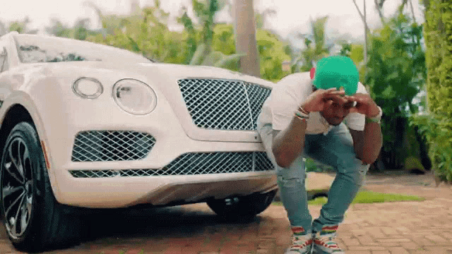 a man wearing a green hat is kneeling in front of a white bentley car .