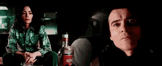 a man and a woman are sitting next to each other in a dark room with a bottle of vodka in the background .