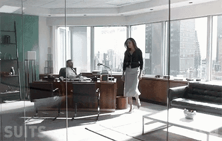 a woman is walking through an office with a man sitting at a desk and a couch .