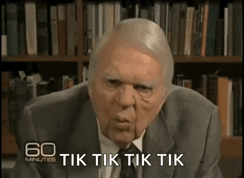 an older man in a suit and tie is sitting in front of a bookshelf and making a funny face .