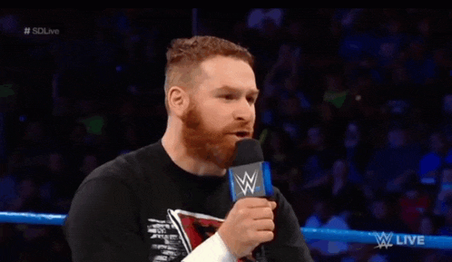 a man with a beard is talking into a microphone while sitting in a wrestling ring .