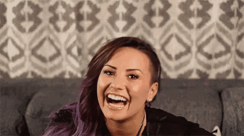 a woman with purple hair is laughing while sitting on a couch in front of a patterned wall .