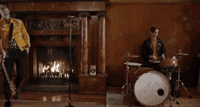 a man in a yellow jacket is singing into a microphone while another man plays drums
