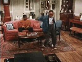 two men are dancing in a living room with a pink couch .