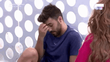 a man is sitting on a bed with his hand on his forehead while a woman looks on .
