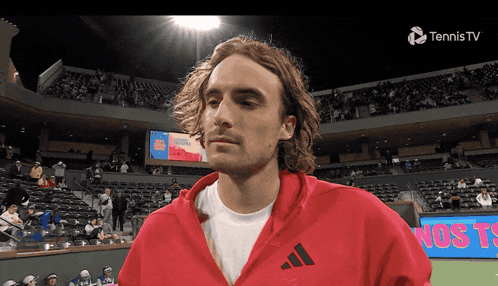 a man wearing a red adidas jacket stands in a tennis stadium