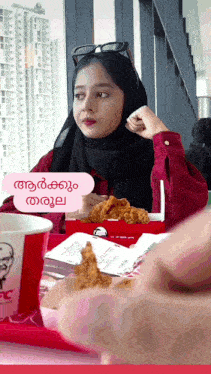 a woman in a hijab is sitting at a table with a box of fried chicken and a kfc cup