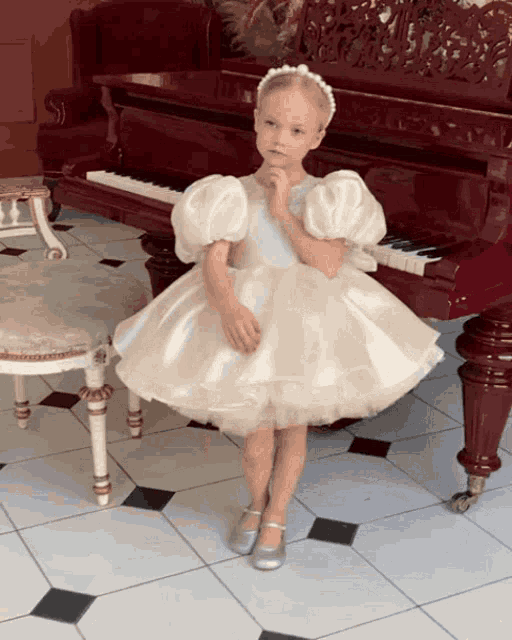 a little girl in a white dress stands in front of a grand piano