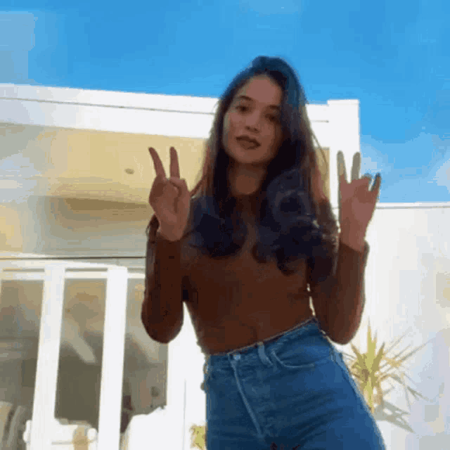 a woman in a brown sweater and blue jeans is giving the peace sign