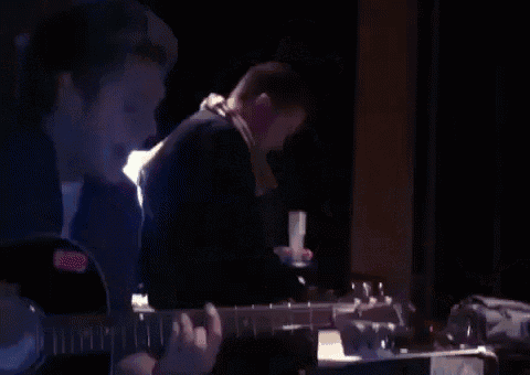 a man is playing a guitar in a dark room while another man looks on