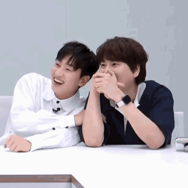 two young men are sitting at a table with their hands covering their faces