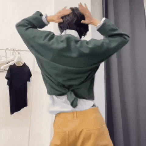 a woman in a green shirt and yellow pants is standing in a dressing room with her hands on her head