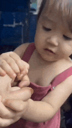 a baby in a pink tank top is holding a person 's hand and looking at it .