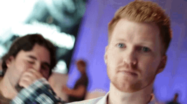 a man with a beard looks at the camera with another man in the background
