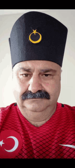 a man with a mustache is wearing a red shirt and a black hat with a crescent moon and star on it