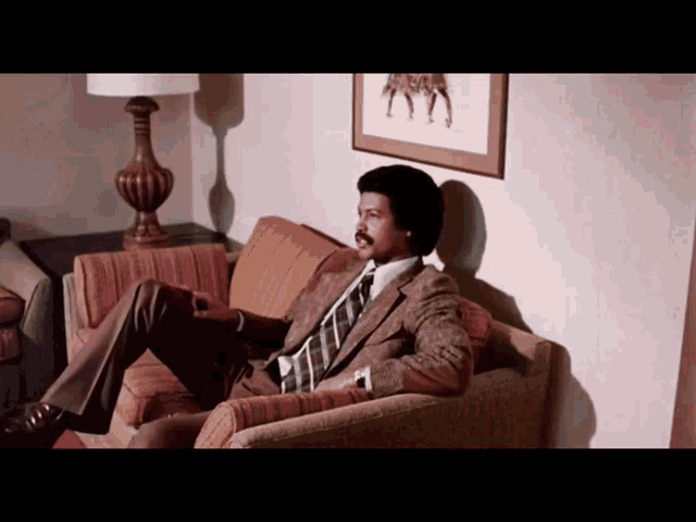 a man in a suit and tie sits on a couch in front of a lamp
