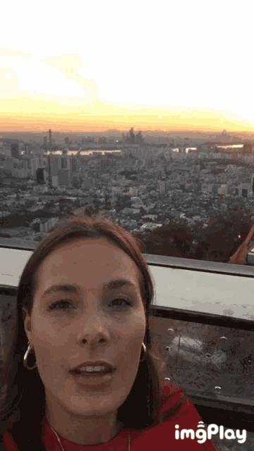 a woman in a red shirt is taking a selfie with a city skyline in the background