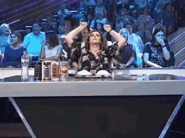 a woman is sitting at a table with her arms in the air in front of a crowd of people