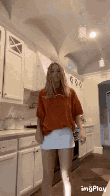 a woman is standing in a kitchen wearing an orange shirt and a white skirt ..