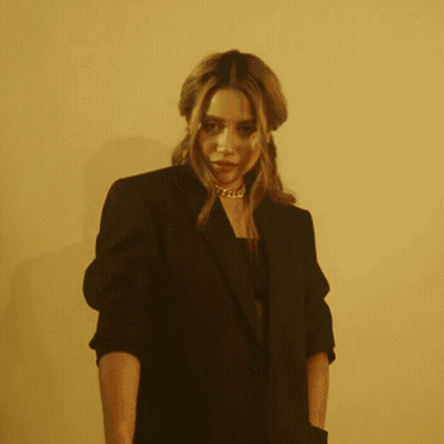 a woman wearing a black blazer and a gold necklace looks at the camera