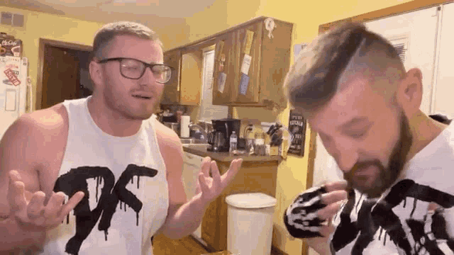 two men are standing next to each other in a kitchen . one of the men is wearing a tank top that says df .