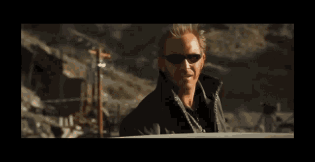 a man wearing sunglasses and a black jacket is standing in front of a mountain .