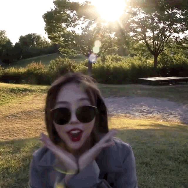 a woman wearing sunglasses stands in a field with the sun shining through the trees in the background