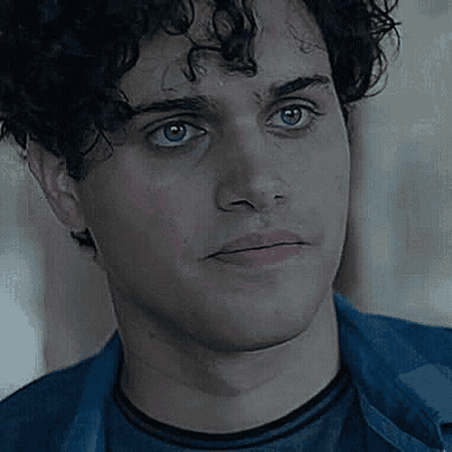 a close up of a man 's face with curly hair wearing a black shirt and a blue jacket .