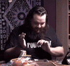a man with a beard wearing a black shirt that says power is sitting at a table eating food .