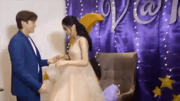 a man is putting a ring on a woman 's finger in front of a purple curtain .