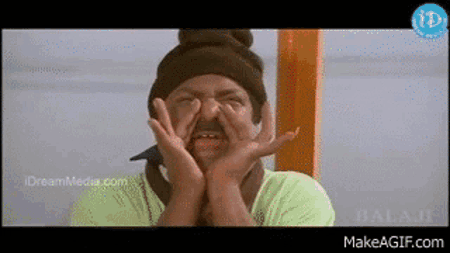 a man wearing a beanie and a green shirt is making a funny face with his hands on his face .