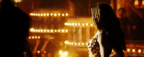 a woman in a gold dress is standing in a dark room with a lot of lights behind her .