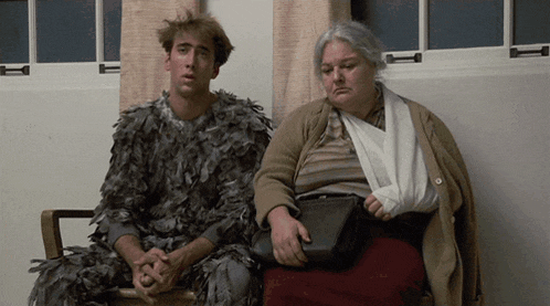 a man dressed in feathers sits next to an older woman with a bandaged arm