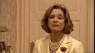 a woman in a yellow suit and pearls is sitting in a chair and making a funny face .